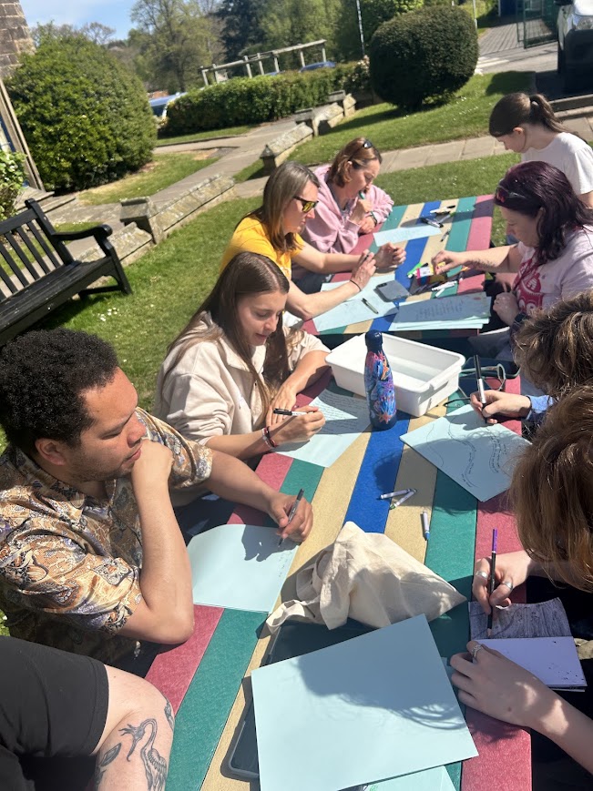 People writing and drawing around an outdoor picnic table.