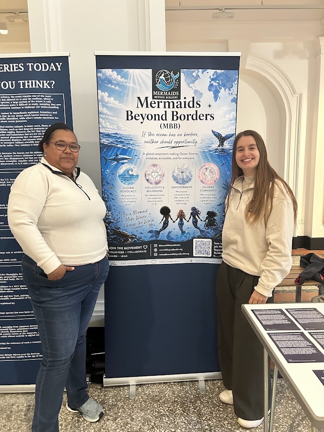 Two people standing beside a Mermaids Beyond Borders banner.