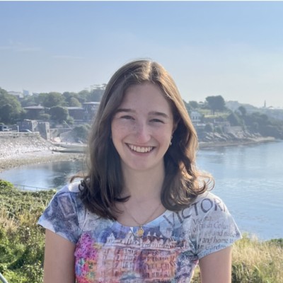Alexandra smiling outdoors by the coast, wearing a patterned top.
